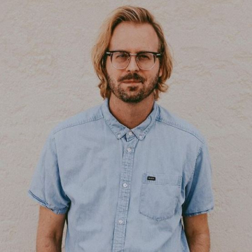 Evan Stowers, hairdresser and owner of The Desert Lounge Salon in St. George, TU wearing a chambray shirt with blonde hair and glasses against a white stucco wall