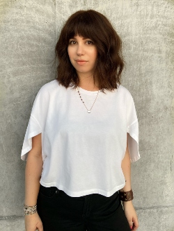 Samantha Sellars, hair stylist and educator, standing in front of a cream stucco wall with dark wavy long bob and bangs, white flowy tshirt and black pants.