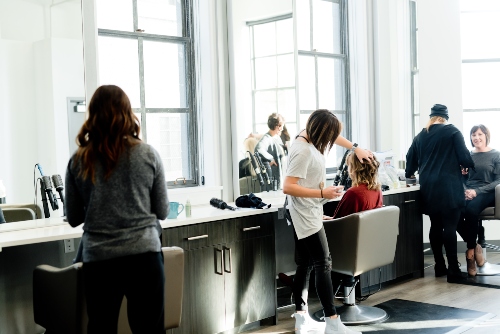 Hair salon with three stylists working on their clients. White walls with high ceilings and large windows letting in a lot of natural light