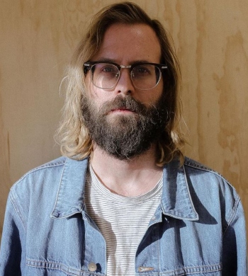 Hairdresser Evan Stowers stands in front of a wood wall with long blonde hair, glasses and a full beard with a stoic look discussing mental health for hair stylists 