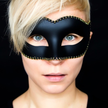 Blond woman with short hair surrounded by a black background and wearing a mask over eyes and nose, with her blue eyes peering out.