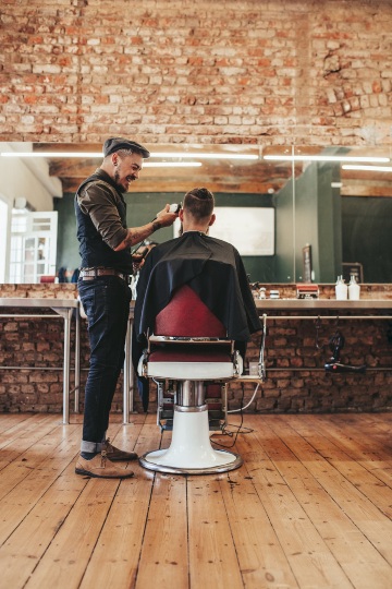 A barber is smiling while cutting his clients hair with clippers in a large open barber shop with wood floors and raw brick walls