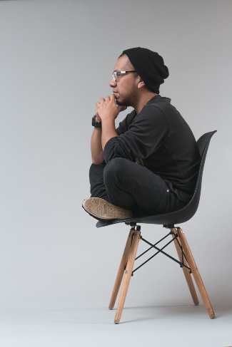 Man in a black slouchy beanie sitting in a black chair with wooden legs. He is wearing glasses, sitting cross legged with his hands on his chin thinking about salon booth or salon suite rental