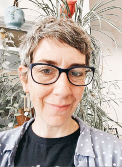 Kate Kent, a woman with salt and pepper pixie cut and dark rimmed glasses stands in front of a plant shelf