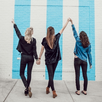 3 young women with their back facing you. they are holding their hands up in the air in front of a brick wall painted with blue and white stripes.