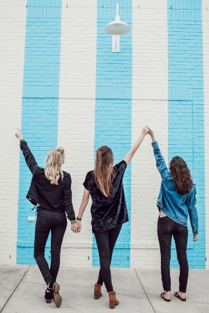 three young girls stand facing a brick wall painted with wide white and blue stripes, arms raised above their heads and ankles crossed