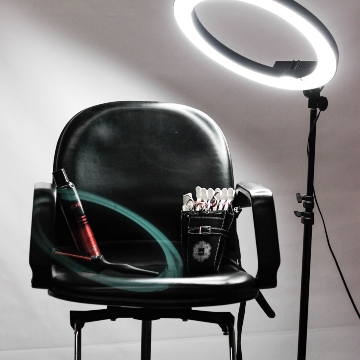 Black salon chair with hairspray, a curling iron and a holster of scissors and clips wits in front of a blank white wall with a ring light shining on it