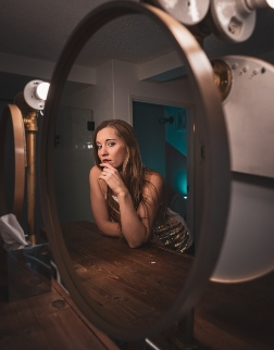 Woman in gold dress with long brown hair stares pensively into a round mirror in a dimly lit salon